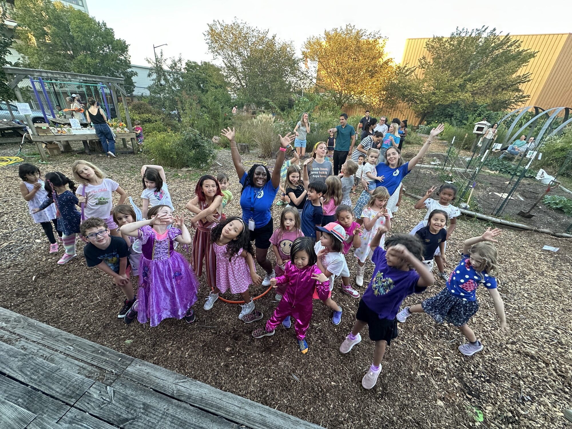 A Janney+ staff member cheering with a big group of kids in the school garden at golden hour