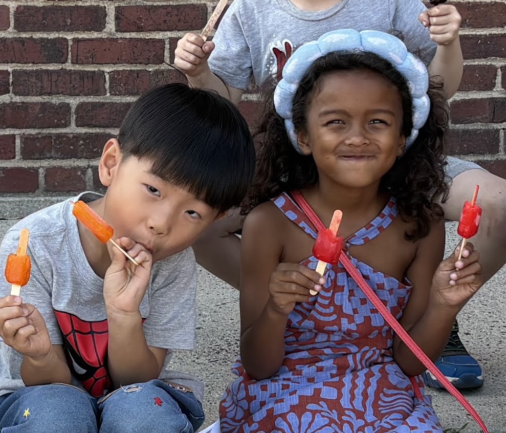 Two students sitting against a brick wall enjoying popsicles on a warm afternoon
